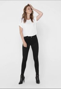 Jeans skinny neri con una superficie liscia, vita alta e dettagli con pannello a ginocchio, abbinati a una maglietta bianca a maniche corte e stivali neri alla caviglia.