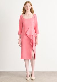 Robe rose mi-longue avec des manches trois-quarts, présentant un détail de volants à l'avant et une fente sur le côté, fabriquée dans un tissu lisse.