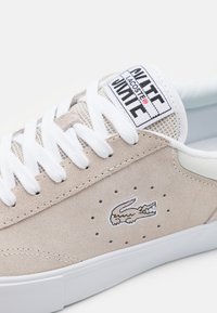 Baskets en daim beige avec lacets blancs et accents en mesh. Dotées de perforations et d'un logo brodé sur le côté, semelle blanche.