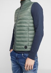 Lättviktsgrön pufferväst med hög krage, horisontell sömnad, frontdragkedja och en liten svart logotyp på fållen.