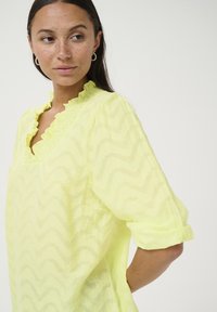 Blusa gialla a fantasia a zigzag con scollatura a V arricciata, maniche lunghe arrotolate e tessuto leggero e texturizzato.