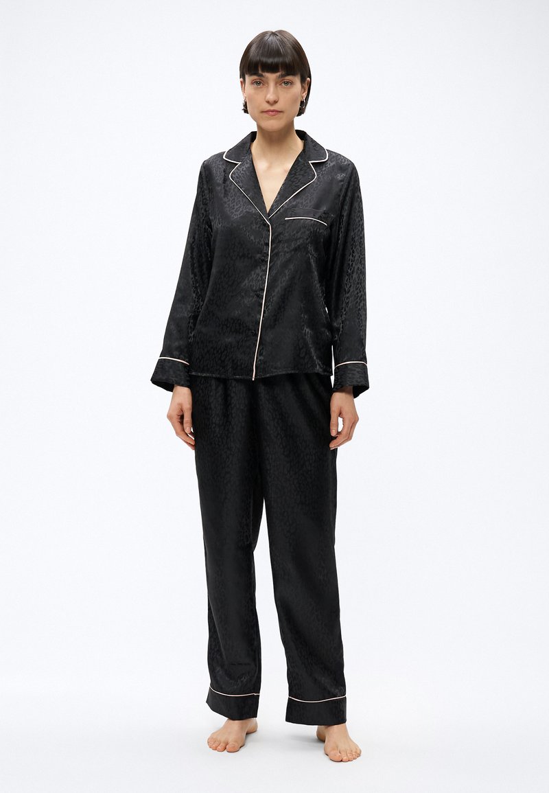 Kobieta stojąca boso, ubrana w czarne satynowe piżamy w panterkę z białą lamówką i długimi rękawami na jednolitym białym tle.