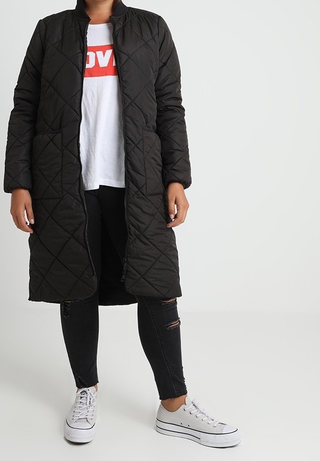 Mustapinta, tikattu pitkä takki, jossa on vetoketju edessä, seisomakaulus ja sivutaskut. Yllä valkoinen T-paita ja mustat revityt farkut, sekä valkoiset lenkkitossut.