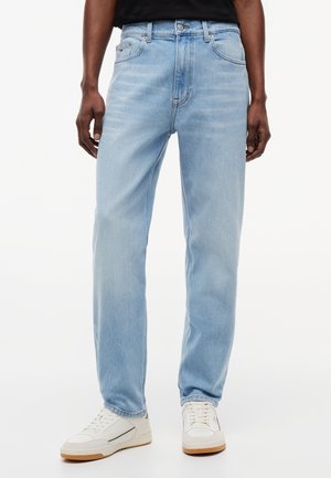 Személy világoskék egyenes szárú farmert és fehér tornacipőt visel, sima fehér háttér előtt áll.