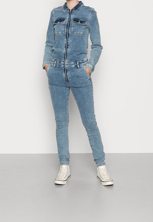 Combinaison en denim bleu clair, avec une fermeture éclair à l'avant, deux poches poitrine et une coupe ajustée. Associée à des baskets montantes blanches.