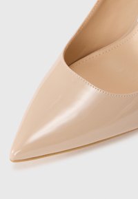 Beige patent leather high-heeled shoe with a pointed toe and smooth surface, featuring a low-cut vamp and minimal stitching detail.
