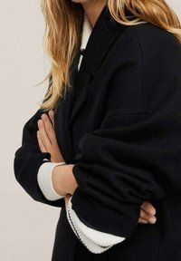 Manteau noir oversize à manches larges, avec des poignets côtelés blancs. Matière douce offrant un aspect texturé. Aucun moyen de fermeture visible.