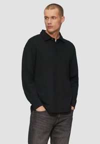 Schwarzes Langarm-Polohemd mit einem kragenloses Ausschnitt und einer zwei knöpfbaren Blende. Kombiniert mit dunkelgrauen Jeans, die eine entspannte Passform zeigen.