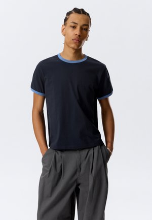 Jeune personne aux cheveux tressés portant un t-shirt noir avec des bordures bleues et un pantalon plissé gris, debout les mains dans les poches.