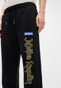 Μαύρες φόρμες με κορδόνι στη μέση, με μπλε λογότυπο "HUGO" και κίτρινο κείμενο "Jujutsu Kaisen" στο κάτω μέρος του ποδιού. Υφασμάτινη επιφάνεια.