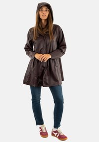 Vrouw in een donkerbruin regenjack met capuchon, blauwe jeans en rood-witte sneakers, staande met de handen gevouwen voor zich.