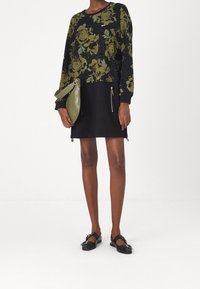 Robe noire à motif floral vert, manches longues, deux poches zippées, assortie à un sac vert et des chaussures plates noires.