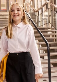 Chemise blanche boutonnée avec col, associée à un pantalon noir taille haute avec poches, tenant un dossier jaune.