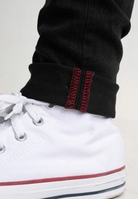 Bílé plátěné boty s kovovými očka a červeným pruhem na podrážce, doplněné o černé džíny s manžetami a červeným prošitím.