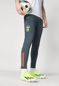 Tamno sivi sportski hlače s narančastim prugama, s logotipom na butu i teksturiranom bočnom panou. Svijetložuti nogometni kopački.