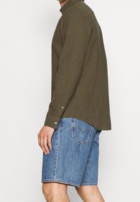 Camisa de manga larga verde oliva, abotonada en los puños, combinada con pantalones cortos de mezclilla azules. La camisa tiene un cuello; los pantalones cortos son hasta las rodillas con costuras visibles.