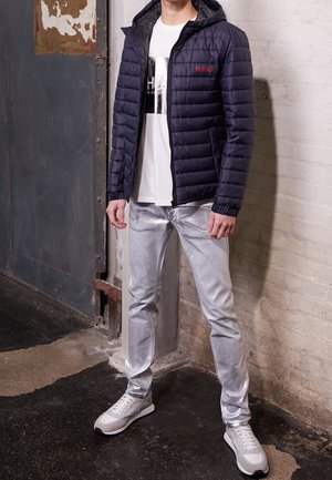Homme portant une veste matelassée bleu marine, un t-shirt blanc à motif, un pantalon argenté brillant et des baskets blanches, debout contre un mur et une porte usés.
