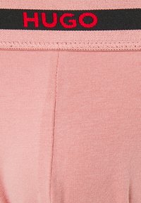 Helles rosa Oberteil aus Baumwollmischung mit einem schwarzen Bund und rotem "HUGO"-Logo. Glatte Textur mit leicht gerippten Details.