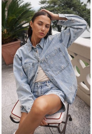 Giacca di jeans azzurro chiaro con ricami floreali, abbinata a un top beige e a pantaloni corti di jeans coordinati. Tessuto morbido, vestibilità rilassata, design casual.