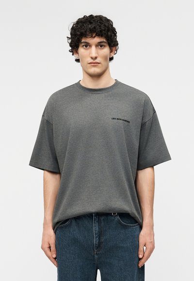 Bărbat cu păr creț purtând un tricou larg gri cu textul „LES BENJAMINS” pe piept și blugi albaștri, stând pe un fundal alb.