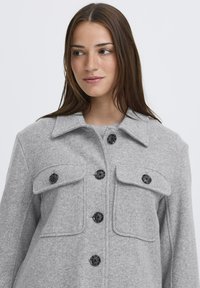 Sivý vlnenný kabát s golierom, ktorý má dve náprsné vrecká a čierne gombíky. Textília má mäkkú štruktúru a perfektný strih.