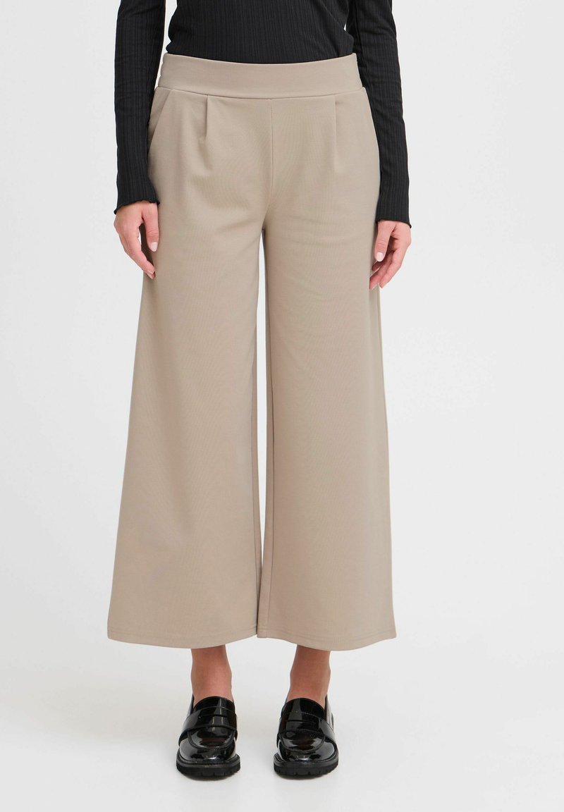 Pantalons larges beiges en tissu doux avec une texture lisse, dotés de plis avant et d'une taille élastique. Des chaussures noires complètent le look.