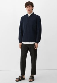 Granatowa bomberka z zamkiem błyskawicznym, ściągaczami przy mankietach i kołnierzu, w połączeniu z czarnymi spodniami slim-fit i czarnymi klapkami.