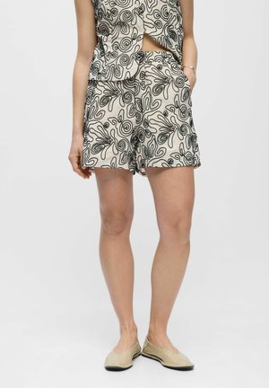 Kvinde iført matchende beige shorts og top med sort abstrakt blomsterbroderi, kombineret med beige flade sko.