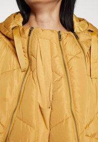 Gul vadderad jacka med en quiltad textur. Har en hög krage, justerbar huva och dubbla dragkedjor framtill.