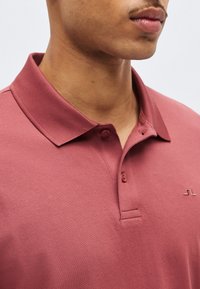 Κοντινό πλάνο ενός ατόμου που φοράει ροζ πόλο πουκάμισο με διπλωμένο γιακά και δύο κουμπιά κουμπωμένα στο λαιμό.