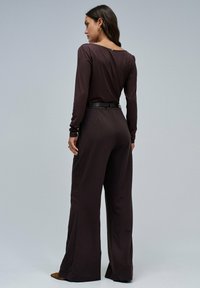 Mono marrón de manga larga que presenta un top ajustado, pantalones de pierna ancha, una cintura con cinturón y un ligero escote en la parte posterior. Textura suave.