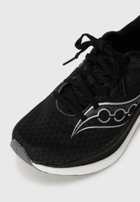 Zapatilla deportiva negra con parte superior de malla texturizada, diseño elegante, cordones planos y una entresuela blanca. Presenta un acento de logotipo plateado en el costado.