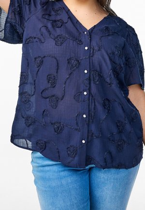 Mujer con una blusa azul marino transparente abotonada con detalles florales texturizados y jeans azul claro, posando frente a un fondo liso.