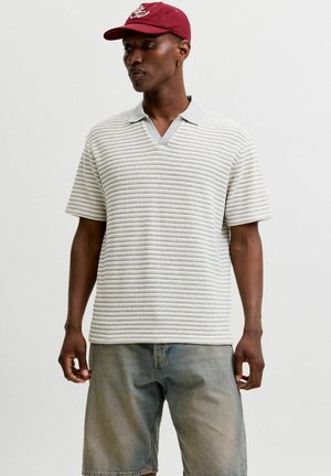 Homme portant un polo gris clair texturé à manches courtes, un short en jean délavé et une casquette de baseball rouge, debout devant un fond uni.