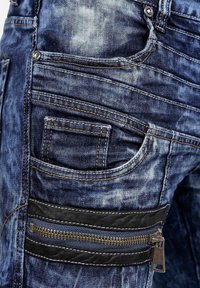 Jeans in denim con finitura blu scuro distressed, cuciture gialle, cinque tasche e un etichetta in pelle nera con dettaglio zipper.