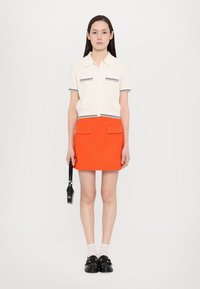 Witte korte mouwen trui met zwarte randen en twee zakken, gecombineerd met een feloranje mini rok met opgestikte zakken, vastgehouden door een zwarte tas.