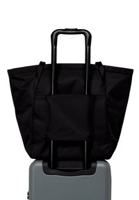Sac fourre-tout noir reposant sur une valise grise, doté d'un design spacieux et de poignées supérieures, avec une poche avant pour un rangement supplémentaire.