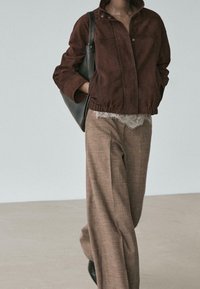 Personne portant une veste en daim marron, un pantalon beige à jambes larges, un haut en dentelle, tenant un sac à épaule noir, debout devant un fond uni.