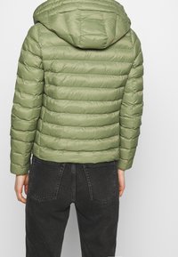 Grøn pufferjakke med hætte, quiltet design, vandret syninger og glat tekstur. Sammen med mørke jeans.