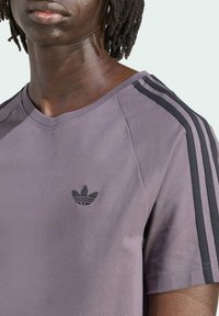 Camiseta de manga corta morada con un logo negro bordado y franjas laterales negras en los hombros. Textura suave, corte casual.