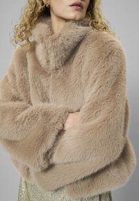 Lažni krzneni šal v svetlo rjavi barvi z visokim ovratnikom, ki ima mehko teksturo in široke rokave, dopolnjuje zlate krilo s sequinami.