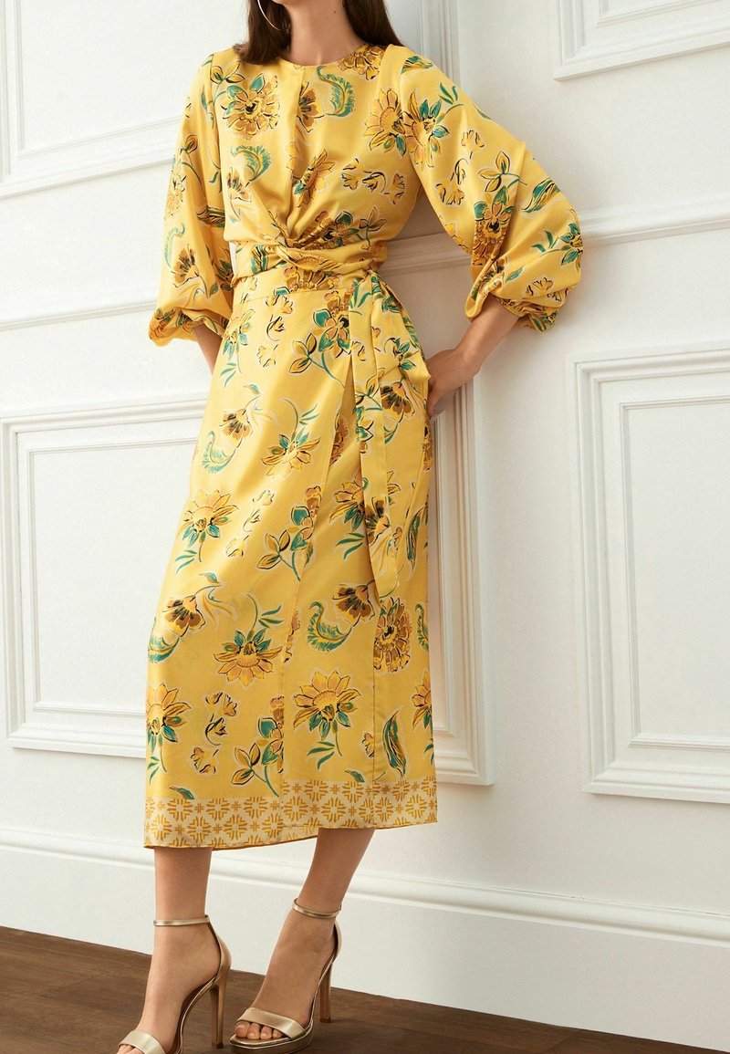 Femme portant une robe midi jaune à fleurs avec manches bouffantes et ceinture à la taille, associée à des sandales dorées à talons hauts, debout près d’un mur blanc.