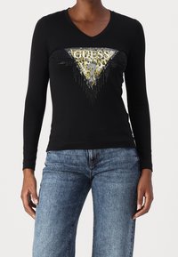 Svart långärmad V-ringad topp med en guld- och djurmönstrad GUESS-logotyptriangel på framsidan, kombinerad med blå denimjeans.
