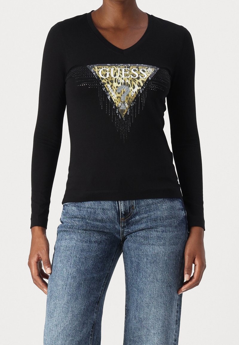 Svart långärmad V-ringad topp med en guld- och djurmönstrad GUESS-logotyptriangel på framsidan, kombinerad med blå denimjeans.