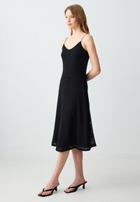 Robe tricotée noire avec un corsage ajusté, une jupe évasée et une texture à motifs. Elle présente des bretelles spaghetti fines et est associée à des sandales noires.