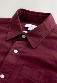 Chemise boutonnée bordeaux avec un col pointu, des boutons blancs et une poche poitrine, étalée à plat montrant l'étiquette du col.