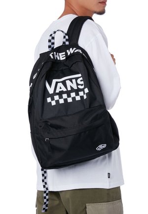 Persona con una mochila Vans negra con patrón de tablero de ajedrez blanco y logo, vestida con camisa blanca y pantalones verde oliva.