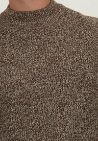 Bruine gebreide trui met een hoge hals, voorzien van een textuurpatroon van afwisselend lichte en donkere strengen voor visueel contrast.