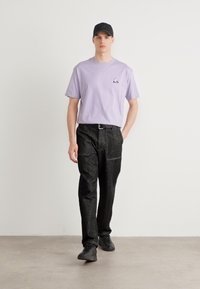 Lilla kortærmet t-shirt med et lille trekantet logo, parret med sorte teksturerede bukser og mørke sneakers, foran en ensfarvet baggrund.