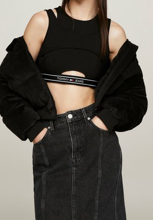 Kobieta nosząca czarny krótki top na ramiączkach z napisem "Tommy Jeans", czarną pikowaną kurtkę opuszczoną na ramiona oraz ciemną spódnicę jeansową z wysokim stanem, z rękami w kieszeniach.